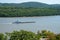 Blue and white tug boat was seen pushing a long barge down the Hudson River. There are small green tree covered hills in the
