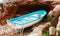 Blue and white wooden rowboat moored between brown rocks and arid sand in front of a cave