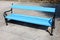 Blue wooden public bench with black wrought iron frame mounted on large concrete tiles sidewalk