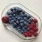 Blueberries rest on a glass dish adorned with a strawberry & raspberries