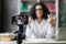 Blur background of pretty multinational woman with dark hair sitting at desk and filming video blog.