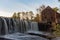 A blur of water flows over stone at dusk by the wooden building and water wheel at the historic Yates Mill County Park in autumn