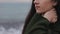 A blurred close view of a beautiful girl in a dark green coat touching her neck at the seashore. The focus shifting from