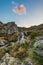 Blurred flowing waterfall in the Alps with colorful sky and clouds at twilight, among rocks, boulders and grass. Little stream in