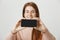 Blurred portrait of cute caucasian redhead model holding smartphone with both hands, advertising it over gray background