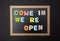 Board with black frame, text Come in, we re open in colorful letters, black wall background