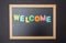 Board with black frame, text Welcome in colorful letters, black wall background