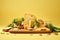 Board with processed cheese, bread and vegetables on pale yellow background