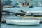 Boats tied up at dock in the rain with marina buildings and sailboats in the background and a couple walking across an entrance