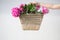 Boho girl holding pink peonies in straw basket. Stylish hipster woman in bohemian floral dress gathering peony flowers on white