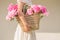 Boho girl holding pink peonies in straw basket. Stylish hipster woman in bohemian floral dress gathering peony flowers on white