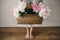 Boho girl holding pink and white peonies in rustic basket and standing barefoot on wooden floor. Stylish hipster woman in bohemian