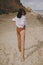 Boho girl in white shirt running on sunny beach, back view. Carefree stylish woman in swimsuit and shirt relaxing on seashore.
