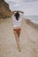 Boho girl in white shirt running on sunny beach, back view. Carefree stylish woman in swimsuit and shirt relaxing on seashore.