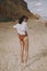 Boho girl in white shirt running on sunny beach, back view. Carefree stylish woman in swimsuit and shirt relaxing on seashore.