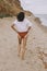 Boho girl in white shirt walking on sunny beach, back view. Carefree stylish woman in swimsuit and shirt relaxing on seashore.
