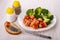 Boiled broccoli with sausages in tomato sauce on plate, fork with sausage on bread, salt and pepper on table