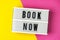 Book now - text on a display lightbox on yellow and pink background