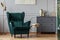 Books on stylish golden small table next to emerald green velvet wing back chair in grey living room interior
