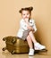Bored baby kid girl with funny buns and in leopard print dress and denim jacket is sitting on modern trunk case, waiting