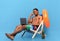 Bored black guy feeling unhappy, having to work online during his summer vacation, sitting in lounge chair with laptop