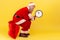 Bored elderly man wearing santa claus costume sitting on big red bag with presents and holding wall clock in hands, waiting time