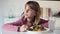 Bored little girl looking sideways while eating a full hamburger with potatoes at home