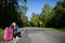 Bored of waiting to hitchhike, a teenager waits at the side of a road leading through a leafy forest on a sunny day.