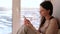 Bored woman watches something on the Internet in her mobile phone while sitting on a windowsill.