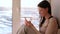 Bored woman watches something on the Internet in her mobile phone and smiling while sitting on a windowsill