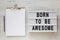 `Born to be awesome` words on a lightbox, clipboard with blank sheet of paper on a white wooden background, top view. Overhead,