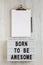 `Born to be awesome` words on a lightbox, clipboard with blank sheet of paper on a white wooden surface, top view. Overhead, fro