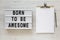 `Born to be awesome` words on a lightbox, clipboard with blank sheet of paper on a white wooden surface, top view. Overhead, fro