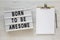 `Born to be awesome` words on a modern board, clipboard with blank sheet of paper on a white wooden background, top view. Overhe