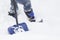 Bottom half of a male in boots and jeans standing in snow with a shovel.