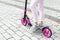 Bottom part of young adult female person wearing pink casual jeans and sneakers riding push kick scooter at city street on summer