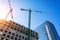 Bottom view of a building under construction with high construction cranes and a glass tower in the background