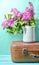 Bouquet of lilac in an old enameled teapot on vintage suitcase on blue background. Retro style still life.