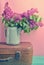 Bouquet of lilac in an old enameled teapot on vintage suitcase on pink background. Retro style still life.