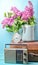 Bouquet of lilacs in enameled kettle on antique suitcase, vintage radio, alarm clock on blue background. Retro style still life.