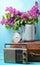 Bouquet of lilacs in enameled kettle on antique suitcase, vintage radio, alarm clock on blue background. Retro style still life.