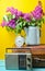 Bouquet of lilacs in enameled kettle on antique suitcase, vintage radio, alarm clock on yellow background. Retro style still life.