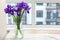 Bouquet of purple irises in a clear glass vase on a linen tablecloth on a wooden table by the window in a modern bright kitchen