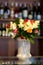 Bouquet of roses in a white ceramic vase counter against the shelf of bottles.