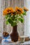 A bouquet of sunflowers in a clay jug and a bowl with cookies on the windowsill behind a lace curtain.