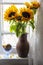 A bouquet of sunflowers in a clay jug and a bowl with cookies on the windowsill behind a lace curtain.
