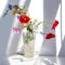 Bouquet of three red poppy flowers and different wildflowers in crystal vase with water on white table with contrast sun light and