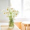 A bouquet of white daisies in a vase on the kitchen table against the background of a window, a refrigerator and stools