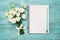 Bouquet of white flowers and empty paper sheet on turquoise rustic table from above. Beautiful vintage card, top view.