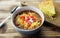 Bowl of chili, with cheese, sour cream, and diced tomatoes in a white bowl on a wooden table, shown with a piece of cornbread.